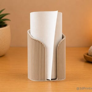 Könnte beinhalten: Ein beigefarbener, zylindrischer Papierrollenhalter mit vertikaler Rippenstruktur und einem U-förmigen Ausschnitt an der Vorderseite. Der Halter enthält eine Rolle weißes Papier. Der Text @3dPrintingLabDE ist unten rechts im Bild zu sehen.