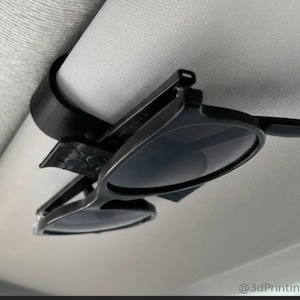 May include: A black 3D-printed plastic clip attached to a light gray vehicle sun visor, designed to hold a pair of black-framed sunglasses securely in place. The clip features a textured surface and the text @3dPrintingLabDE in the corner.