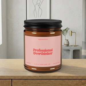 May include: A brown glass jar candle with a black lid. The label is pink with the words "Professional Overthinker" in red. The candle is handmade and has a burn time of 40 to 60 hours.