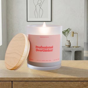 May include: A lit, frosted glass candle with a wooden lid. The candle has a pink label with the words "Professional Overthinker" in red. The candle is on a wooden surface.