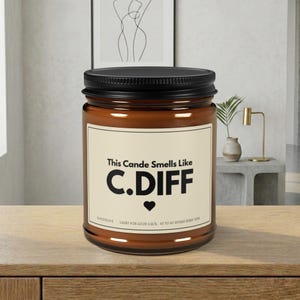 May include: A brown glass jar candle with a black lid. The label reads "This Cande Smells Like C.DIFF" with a heart symbol. The text "HANDMADE LIGHT FOR GOOD LUCK 40 TO 60 HOURS BURN TIME" is also visible.