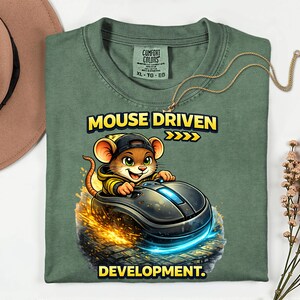 May include: Green t-shirt featuring the text "MOUSE DRIVEN DEVELOPMENT" and a cartoon mouse driving a computer mouse. The mouse wears a black cap and yellow jacket. A gold necklace is draped across the shirt.