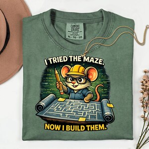 May include: Green Comfort Colors t-shirt featuring a cartoon mouse in a hard hat and glasses, holding a pencil, and looking at a maze blueprint. The text on the shirt reads "I TRIED THE MAZE. NOW I BUILD THEM."