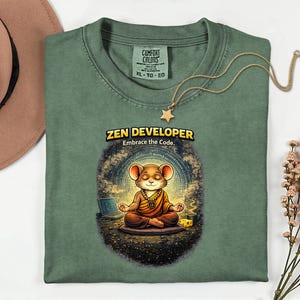May include: Sage green t-shirt featuring a meditating mouse graphic, a laptop, and the text "ZEN DEVELOPER. Embrace the Code." A gold necklace with a star pendant is on the shirt. The shirt is labelled XL-TG-EG.