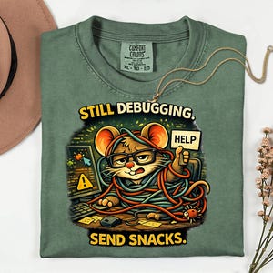 May include: Green Comfort Colors t-shirt featuring a cartoon mouse in glasses, tangled in wires, holding a "HELP" sign. Text reads "STILL DEBUGGING. SEND SNACKS."
