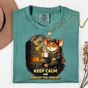 Op de afbeelding: Teal Comfort Colors T-shirt met een cartoonmuis in een hoodie met een kopje koffie, met de tekst "KEEP CALM AND REBOOT THE SERVER." Een gouden sterrenketting is over het shirt gedrapeerd. Een bruine hoed staat in de linkerbovenhoek.