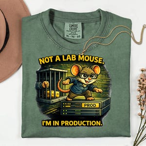 Może przedstawiać: Zielona koszulka z napisem "NOT A LAB MOUSE. I'M IN PRODUCTION." przedstawia mysz z kreskówki w okularach i niebieskiej koszuli. Mysz stoi na stosie sprzętu elektronicznego. Złoty naszyjnik jest zarzucony na koszulę.