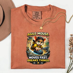 May include: A rust-coloured t-shirt featuring the text "AGILE MOUSE. MOVES FAST. BREAKS THINGS." and a cartoon mouse graphic. The mouse is wearing a cap and jacket, using a computer mouse. A gold necklace with a star pendant is draped on the shirt.