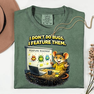 May include: Green Comfort Colors t-shirt featuring the text "I DON'T DO BUGS. I FEATURE THEM." and a cartoon mouse. The mouse is wearing a yellow hoodie and sitting at a desk with a laptop and a whiteboard labeled "FEATURE ROADMAP".