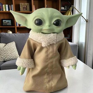 K&ouml;nnte beinhalten: Eine gr&uuml;n geh&auml;utete Puppe mit gro&szlig;en schwarzen Augen und spitzen Ohren, bekleidet mit einem beigefarbenen Gewand mit wei&szlig;em, flauschigem Besatz. Die Puppe ist eine Figur aus einem beliebten Science-Fiction-Franchise.