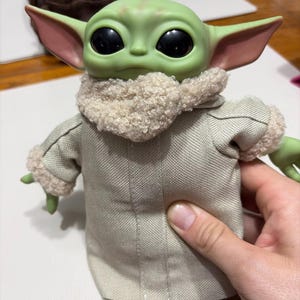 K&ouml;nnte beinhalten: Eine gr&uuml;n geh&auml;utete Puppe mit gro&szlig;en schwarzen Augen und gro&szlig;en Ohren. Die Puppe tr&auml;gt einen beigefarbenen Mantel mit flauschigem Kragen und Manschetten. Die Puppe ist eine Figur aus einem beliebten Science-Fiction-Franchise.