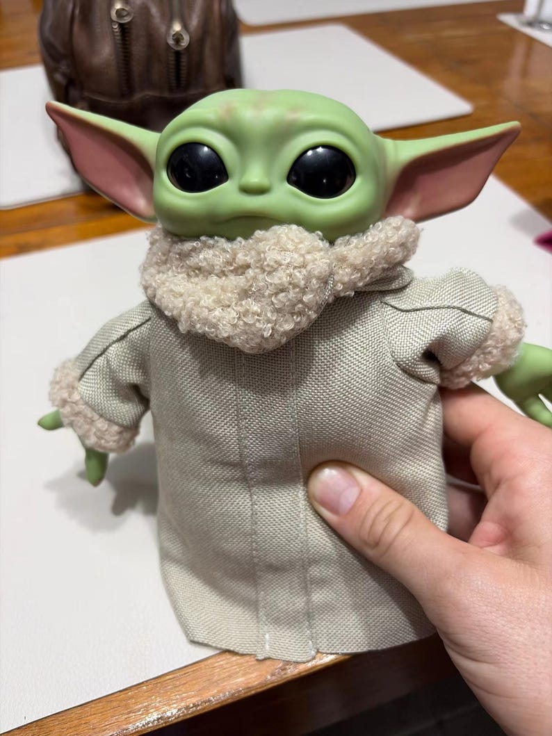 K&ouml;nnte beinhalten: Eine gr&uuml;n geh&auml;utete Puppe mit gro&szlig;en schwarzen Augen und gro&szlig;en Ohren. Die Puppe tr&auml;gt einen beigefarbenen Mantel mit flauschigem Kragen und Manschetten. Die Puppe ist eine Figur aus einem beliebten Science-Fiction-Franchise.