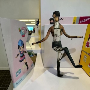 Puede incluir: Una figura recortada de papel de un personaje de la serie de anime Attack on Titan, posando con una pierna levantada. La figura lleva una chaqueta marrón, una camisa blanca, pantalones negros y botas. Una colorida exhibición con personajes de anime está en el fondo.
