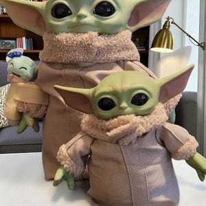 K&ouml;nnte beinhalten: Drei Pl&uuml;schpuppen von Grogu in verschiedenen Gr&ouml;&szlig;en werden gezeigt. Die gr&ouml;&szlig;te Puppe befindet sich in der Mitte, mit einer kleineren links und einer mittelgro&szlig;en davor. Jede Puppe hat gro&szlig;e schwarze Augen, gr&uuml;ne Haut und beige Roben.