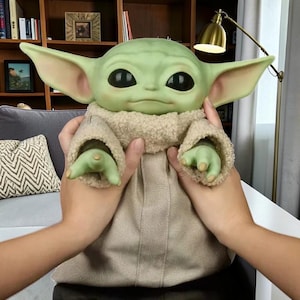 K&ouml;nnte beinhalten: Eine gr&uuml;nh&auml;utige Puppe mit gro&szlig;en Ohren und schwarzen Augen, die einen beigefarbenen Umhang mit flauschigem Kragen tr&auml;gt. Die Puppe wird von zwei H&auml;nden gehalten, die ihre detaillierten Merkmale und ihr Design hervorheben.