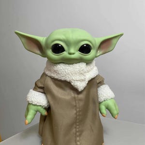 K&ouml;nnte beinhalten: Eine gr&uuml;n geh&auml;utete Puppe mit gro&szlig;en schwarzen Augen und &uuml;bergro&szlig;en Ohren. Die Puppe tr&auml;gt einen beigefarbenen Mantel mit einem wei&szlig;en, flauschigen Kragen und Manschetten. Die H&auml;nde und F&uuml;&szlig;e der Puppe sind gr&uuml;n mit orangefarbenen N&auml;geln. Die Puppe ist eine Figur aus einem beliebten Science-Fiction-Franchise.