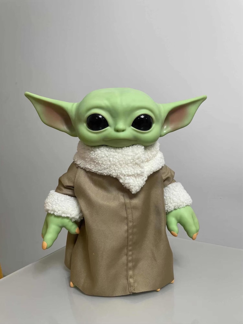 K&ouml;nnte beinhalten: Eine gr&uuml;n geh&auml;utete Puppe mit gro&szlig;en schwarzen Augen und &uuml;bergro&szlig;en Ohren. Die Puppe tr&auml;gt einen beigefarbenen Mantel mit einem wei&szlig;en, flauschigen Kragen und Manschetten. Die H&auml;nde und F&uuml;&szlig;e der Puppe sind gr&uuml;n mit orangefarbenen N&auml;geln. Die Puppe ist eine Figur aus einem beliebten Science-Fiction-Franchise.