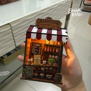 Könnte beinhalten: Ein Miniaturmodell eines Cafés mit einer rot-weiß gestreiften Markise. Das Schild lautet "GRANDSTAND COFFEE CO. AROMA & BREW." Eine Tafel zeigt die Kaffeepreise. Das Modell enthält Regale mit Miniatur-Kaffeeartikeln.