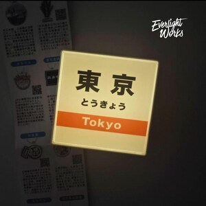 May include: A square, illuminated sign featuring the Japanese kanji for Tokyo, the hiragana characters for Tokyo, and the English word Tokyo in white text on an orange horizontal band. The sign emits a soft, warm glow against a dark background.
