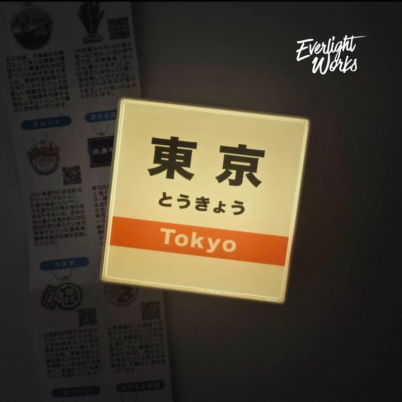 May include: A square, illuminated sign featuring the Japanese kanji for Tokyo, the hiragana characters for Tokyo, and the English word Tokyo in white text on an orange horizontal band. The sign emits a soft, warm glow against a dark background.