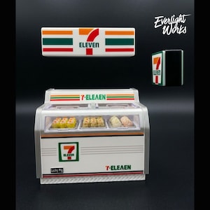 May include: Miniature 7-Eleven convenience store display with a white, orange, green, and red color scheme. The display case contains small food items. The 7-Eleven logo is visible on the display and on a separate sign.