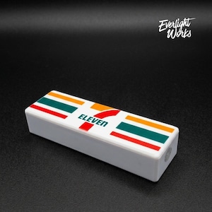May include: A white rectangular object with the 7-Eleven logo on top. The logo features the word "ELEVEN" in red and green, orange, and red stripes. The object is on a black surface.