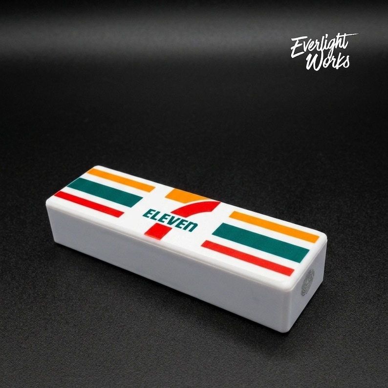 May include: A white rectangular object with the 7-Eleven logo on top. The logo features the word "ELEVEN" in red and green, orange, and red stripes. The object is on a black surface.