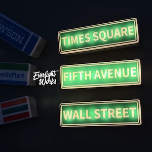 Pode incluir: Três placas retangulares iluminadas com fundo verde e bordas brancas, exibindo o texto TIMES SQUARE, FIFTH AVENUE e WALL STREET. À esquerda, três placas retangulares menores apresentam os logotipos da Lawson, FamilyMart e 7-Eleven.