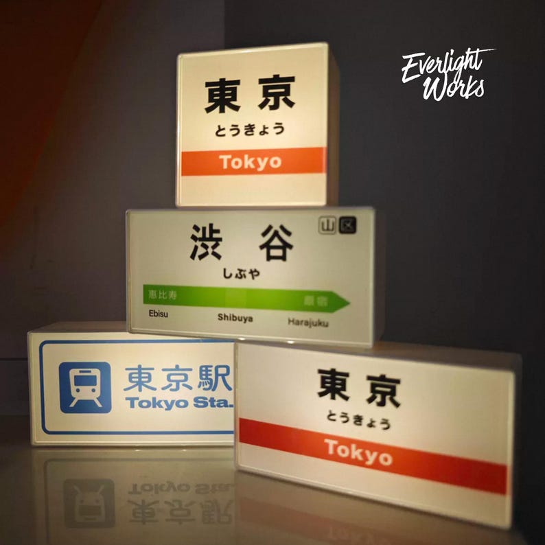May include: Four rectangular illuminated signs featuring Japanese train station designs. One sign displays Tokyo in Japanese and English with an orange stripe. Another shows Shibuya with a green arrow. A third sign features the Tokyo Station logo.