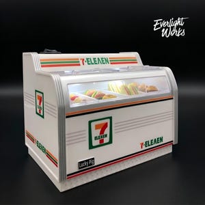 May include: A miniature 7-Eleven display case with a white and silver finish. The case features the 7-Eleven logo and contains miniature sandwiches. The text "7-ELEVEN" is displayed on the case.