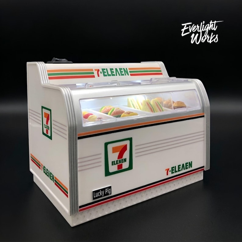 May include: A miniature 7-Eleven display case with a white and silver finish. The case features the 7-Eleven logo and contains miniature sandwiches. The text "7-ELEVEN" is displayed on the case.
