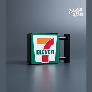 May include: A miniature 7-Eleven store sign with a green border, white background, and the number seven in orange and red. The word "ELEVEN" is in green. The sign is mounted on a black bracket. The text "Everlight Works" is in the upper right corner.