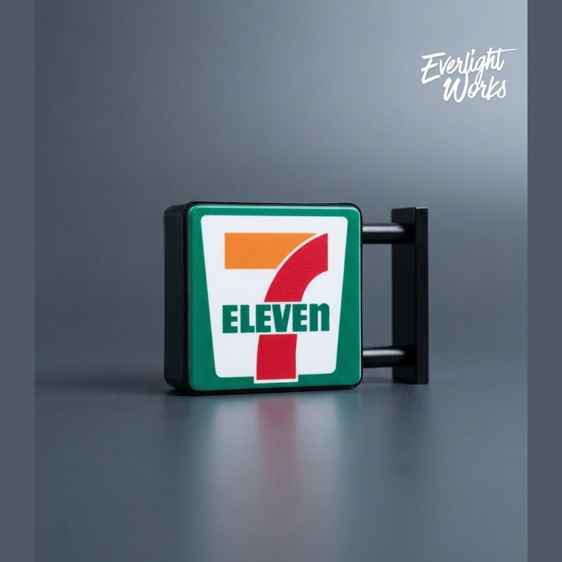 May include: A miniature 7-Eleven store sign with a green border, white background, and the number seven in orange and red. The word "ELEVEN" is in green. The sign is mounted on a black bracket. The text "Everlight Works" is in the upper right corner.