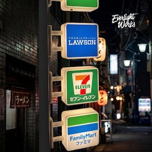 Può includere: Una pila verticale di tre insegne quadrate illuminate con i loghi di Lawson, 7-Eleven e FamilyMart. Ogni insegna è montata su un palo bianco e mostra i nomi dei marchi in inglese e giapponese sui loro colori caratteristici.