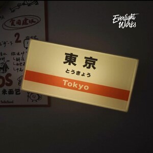 May include: A rectangular illuminated sign featuring the Japanese kanji for Tokyo, the hiragana characters, and the English word Tokyo printed in white on an orange horizontal stripe. The sign has a cream-colored background and a thin border.