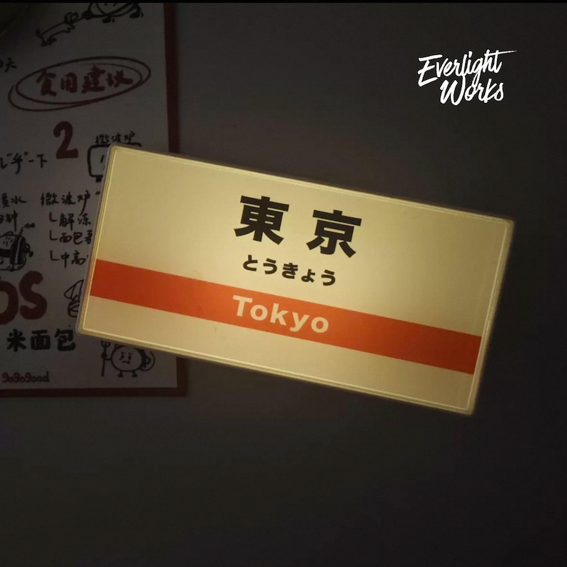 May include: A rectangular illuminated sign featuring the Japanese kanji for Tokyo, the hiragana characters, and the English word Tokyo printed in white on an orange horizontal stripe. The sign has a cream-colored background and a thin border.