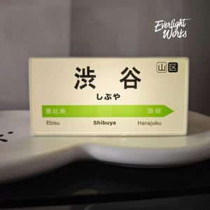 May include: A rectangular, off-white illuminated sign featuring the Japanese text for Shibuya, with a green horizontal arrow pointing right. The sign displays the station names Ebisu, Shibuya, and Harajuku in both Japanese and English text.