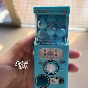 以下が含まれることがあります： COSMIC CODEと書かれた水色のミニチュア自動販売機のおもちゃ。上部の透明なケースには小さな青い球体が入っており、中央には白いダイヤル、下部には円形の取り出し口と文字がプリントされています。