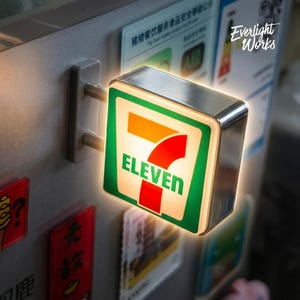 Könnte beinhalten: Ein quadratisches, beleuchtetes 7-Eleven-Wandschild mit einem Rahmen aus gebürstetem Metall. Das Schild zeigt das ikonische 7-Eleven-Logo mit grünem Hintergrund, weißem Rand, der Zahl 7 in Orange und Rot sowie dem Wort ELEVEN in Grün.