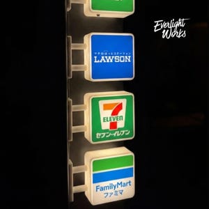 以下が含まれることがあります： Lawson、7-Eleven、FamilyMartのロゴが入った4つの正方形の照明付き看板が縦に並んでいます。各看板は白いブラケットが付いた中央の黒い柱に取り付けられています。右上にEverlight Worksというテキストが表示されています。