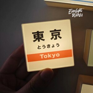 May include: A square, illuminated sign featuring the Japanese kanji for Tokyo, the hiragana characters for Tokyo, and the English word Tokyo printed in white on an orange horizontal band. The sign has a white background and a thin border.