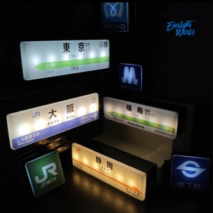 May include: A collection of illuminated rectangular and square signs featuring Japanese train station names and transit logos. The signs display text in Japanese characters and English, including Tokyo, Osaka, Fukushima, and Shizuoka, with bright LED backlighting against a dark background.