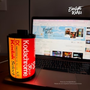 May include: A cylindrical lamp shaped like a vintage Kodak Kodachrome 64 film canister with a glowing yellow and red exterior. The label features the text Kodak Select, DX Process K-14 36-64 KR, and Kodachrome 64 36 EXP. It sits on a laptop.