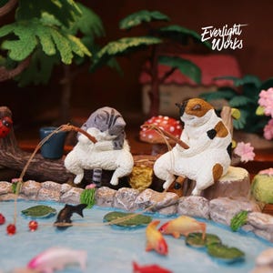 Può includere: Due statuette di gatti in miniatura, uno tigrato grigio e uno calico, seduti su un tronco a pescare in uno stagno blu con ninfee e pesci colorati. La scena include un fungo rosso, alberi verdi e il testo Everlight Works nell'angolo in alto.