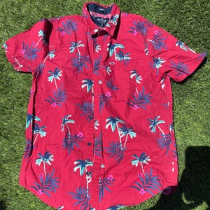 May include: Red short-sleeve button-down shirt with a tropical palm tree pattern in blue, white, and pink. The shirt has a collared neckline and a single chest pocket. The brand Hollister is visible on the shirt.