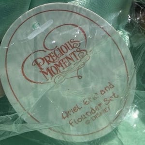 May include: A round tag with the words "Precious Moments" in a cursive font, with the text "Ariel, Eric and Flounder Set" and the Disney logo. The tag is in a clear plastic wrapper.
