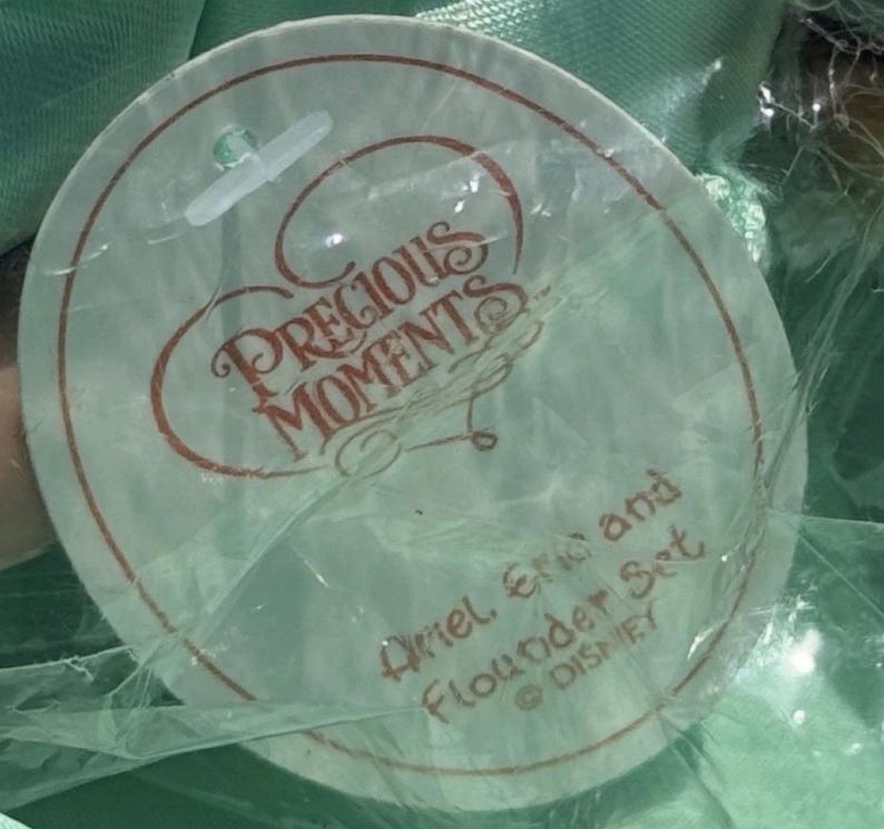 May include: A round tag with the words "Precious Moments" in a cursive font, with the text "Ariel, Eric and Flounder Set" and the Disney logo. The tag is in a clear plastic wrapper.