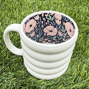 May include: A white ceramic mug with a textured, ridged exterior and a speckled finish. The interior features a floral pattern with pink flowers, green leaves, and a dark background. The mug has a curved handle.