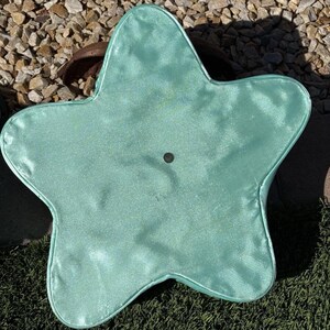 May include: A light teal, star-shaped pillow with a shiny, satin-like texture. The pillow has a small, dark button in the center and is placed on a surface of green turf and small rocks.