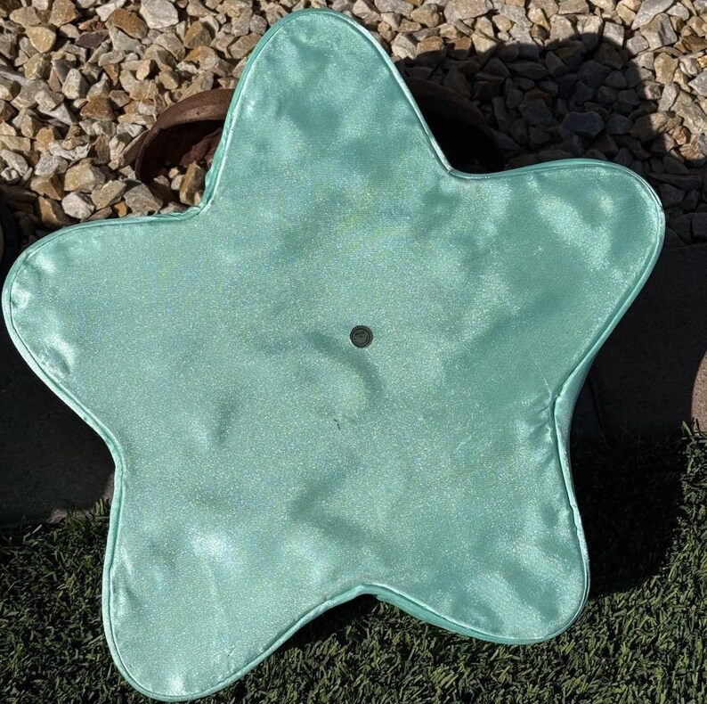 May include: A light teal, star-shaped pillow with a shiny, satin-like texture. The pillow has a small, dark button in the center and is placed on a surface of green turf and small rocks.